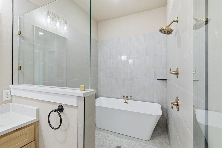 Bathroom featuring a freestanding tub, glass-enclosed shower, gold-toned plumbing fixtures, light wood-finish cabinetry, and a vanity with a white countertop
