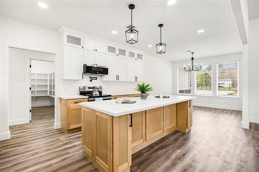 This modern kitchen features a spacious island with light wood cabinetry and a sleek white countertop. It opens to a bright, airy dining area with large windows overlooking a scenic view. The space includes elegant pendant lighting and a convenient pantry.