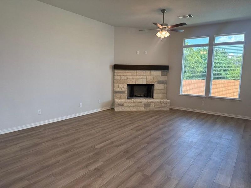Spacious living area featuring wood-finish flooring, a stone fireplace with a substantial wood mantel, multiple large windows with blinds, and a ceiling fan Spacious living area featuring wood-finish flooring, a stone fireplace with a substantial wood mantel, multiple large windows with blinds, and a ceiling fan