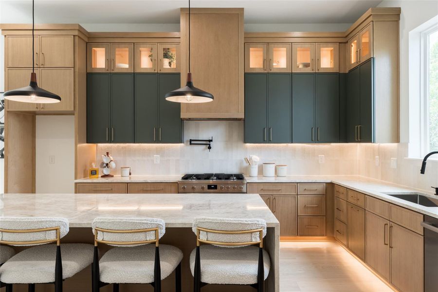 Close-up call sheet: a full-height custom wood range hood with a curved reveal at the base, stacked oak-and-green cabinetry with glass-front display uppers, brass cabinet pulls, a matte-black pot filler, a pro-style six-burner gas range, a honed stone backsplash, and continuous under-cabinet plus toe-kick LED lighting.