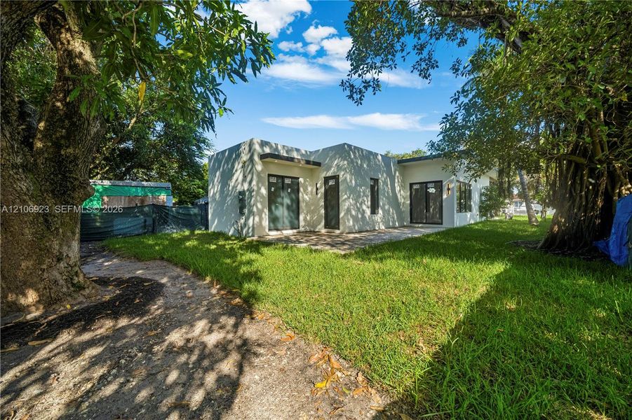 Stucco exterior residence featuring a flat roofline, glass sliding doors, dark-frame windows, and an expansive grass yard with mature trees Stucco exterior residence featuring a flat roofline, glass sliding doors, dark-frame windows, and an expansive grass yard with mature trees