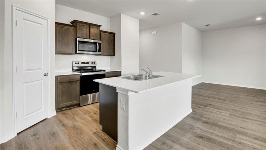 Modern kitchen featuring a central island with a double basin sink, white countertops, stainless steel appliances, dark wood-finish cabinetry, and wood-finish flooring