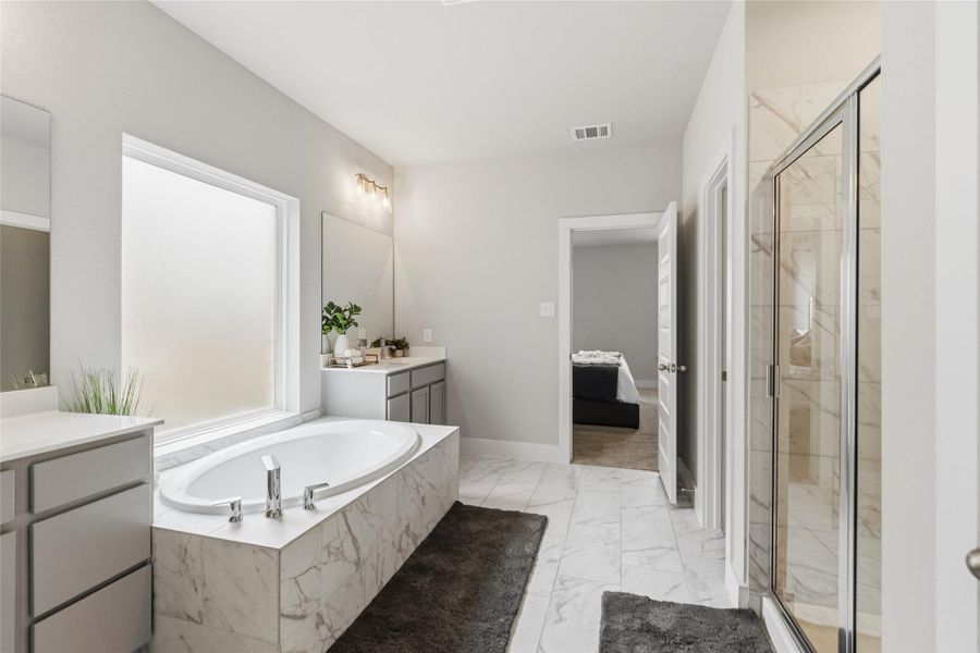 Unwind in your luxurious primary bathroom featuring a soaking tub, separate glass-enclosed shower, dual vanities, and elegant marble-look tile finishes designed for everyday comfort and relaxation.