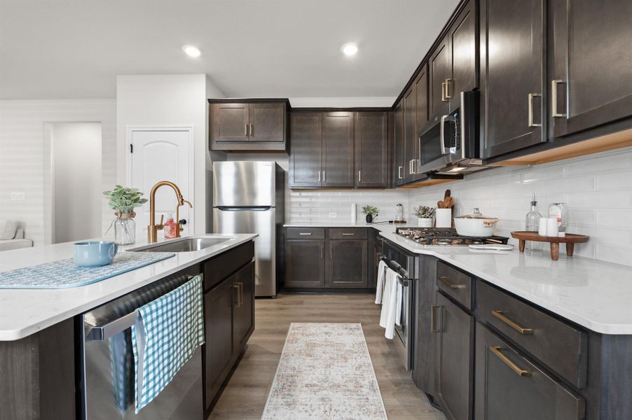 Kitchen featuring dark brown cabinets, appliances with stainless steel finishes, light stone counters, an island with sink, and light wood-style floors Kitchen featuring dark brown cabinets, appliances with stainless steel finishes, light stone counters, an island with sink, and light wood-style floors