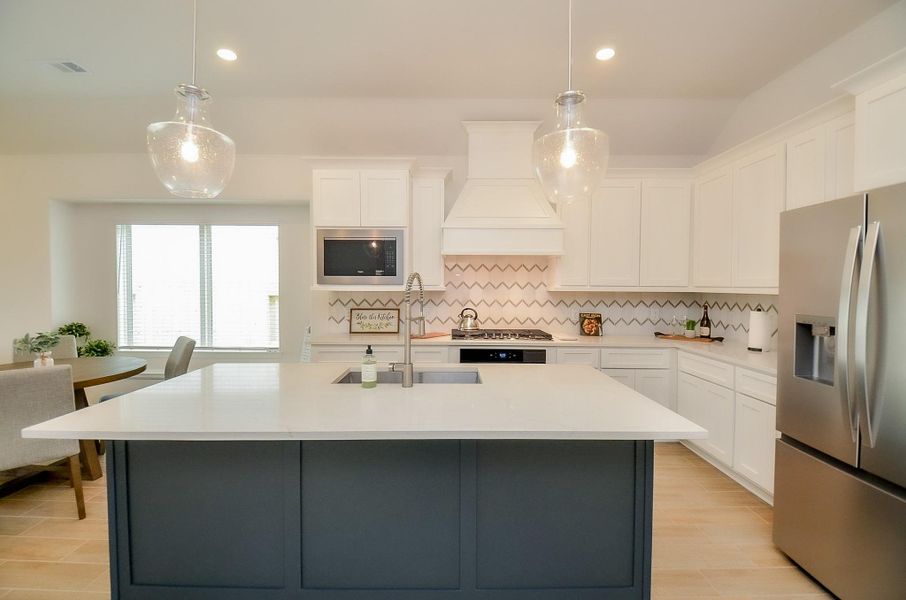 This modern kitchen features a spacious island with a sleek quartz countertop, stainless steel appliances, white cabinetry, and stylish pendant lighting. The herringbone backsplash adds a touch of elegance, and the open layout enhances the bright, inviting atmosphere.