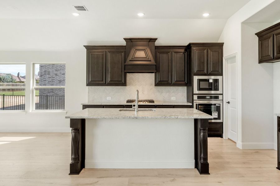 Kitchen with dark wood cabinets, granite island, herringbone tile backsplash, and stainless steel appliances on light hardwood floors