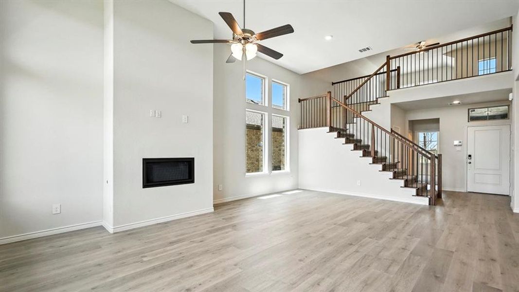 Two-story living area featuring a built-in fireplace, wood-finish flooring, a contemporary ceiling fan, and a prominent staircase with dark wood handrails and black metal balusters