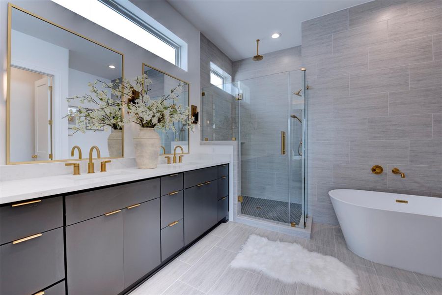 This modern bathroom features a sleek glass-enclosed shower with gold fixtures, a freestanding bathtub, light gray tiles, and elegant decor, offering a luxurious and serene atmosphere.