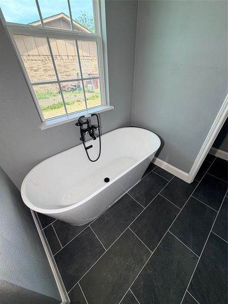 Freestanding soaking tub with a floor-mounted matte black faucet, dark rectangular floor tiling, and a white framed window