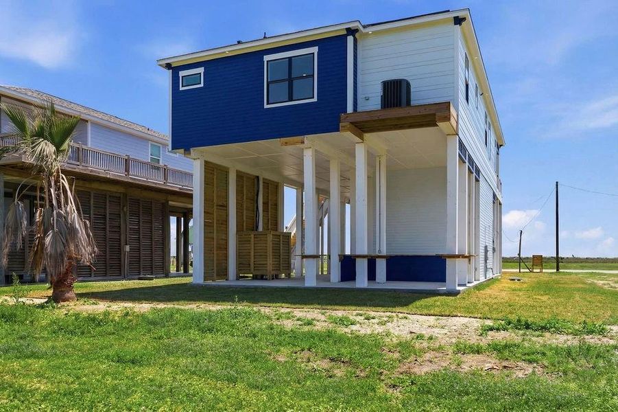 This elevated coastal home features a modern two-story design with a vibrant blue and white exterior. It includes an open ground level ideal for parking or storage, set on a spacious grassy lot with palm trees and ocean views in the distance.