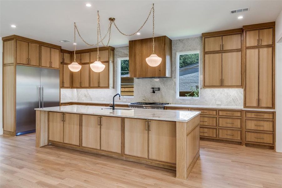 Kitchen with light stone countertops, stainless steel built in fridge, decorative backsplash, decorative light fixtures, and light wood-type flooring Kitchen with light stone countertops, stainless steel built in fridge, decorative backsplash, decorative light fixtures, and light wood-type flooring