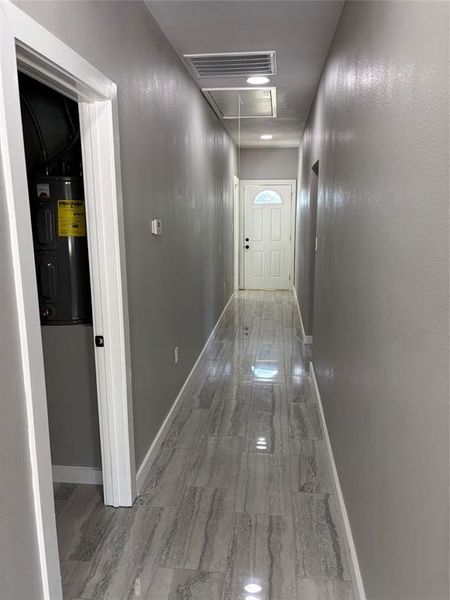 Hallway featuring attic access, water heater, and light marble finish flooring Hallway featuring attic access, water heater, and light marble finish flooring