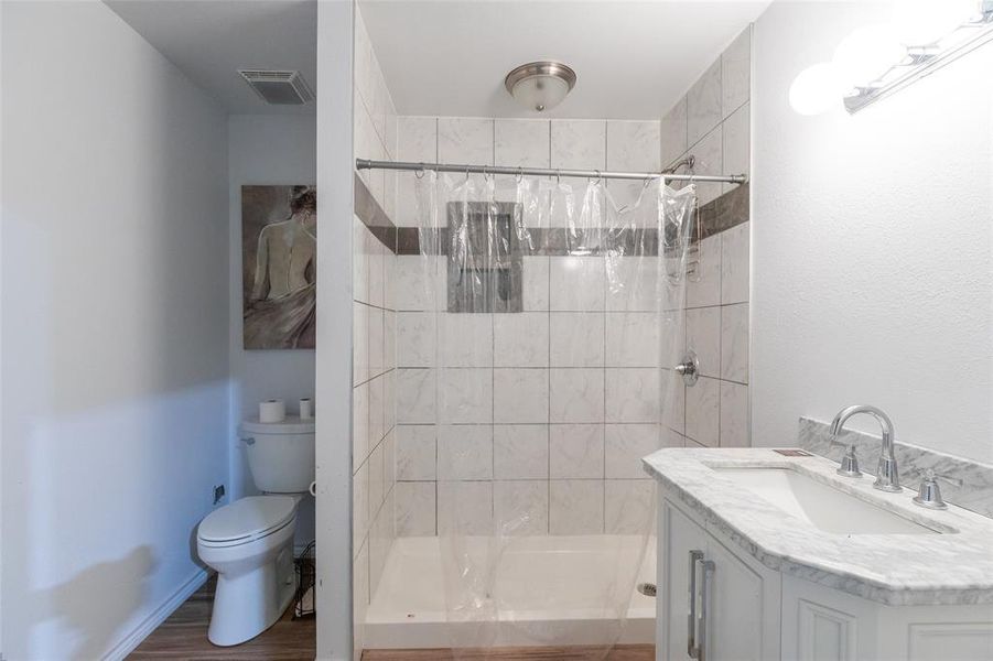 Full bathroom featuring a white vanity with a marble-finish countertop and undermount sink, chrome faucet, tiled shower with a niche, and wood-finish flooring Full bathroom featuring a white vanity with a marble-finish countertop and undermount sink, chrome faucet, tiled shower with a niche, and wood-finish flooring