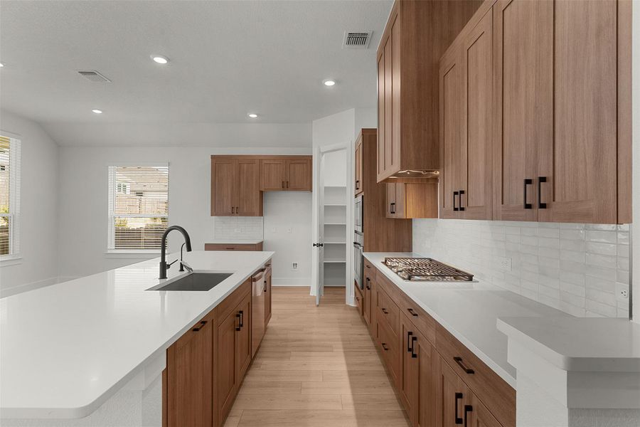 Kitchen featuring a large central island with an undermount sink and matte black faucet, extensive wood-finish cabinetry with black hardware, white countertops, and a white subway tile backsplash Kitchen featuring a large central island with an undermount sink and matte black faucet, extensive wood-finish cabinetry with black hardware, white countertops, and a white subway tile backsplash