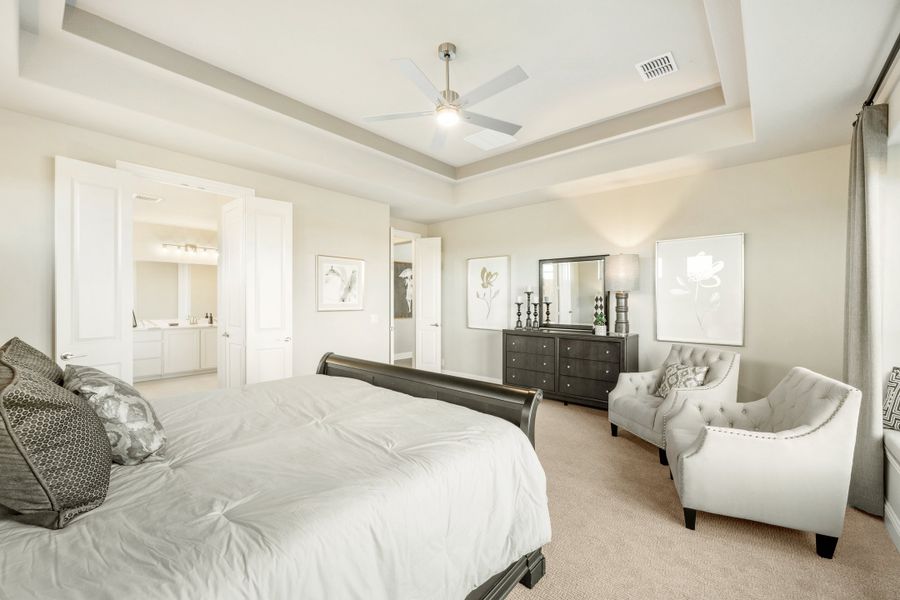 Master bedroom with tray ceiling, ceiling fan, seating area, and ensuite bathroom access Master bedroom with tray ceiling, ceiling fan, seating area, and ensuite bathroom access