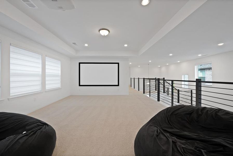 Spacious loft area featuring a mounted projector and screen, recessed lighting, neutral carpeting, and a modern black metal railing