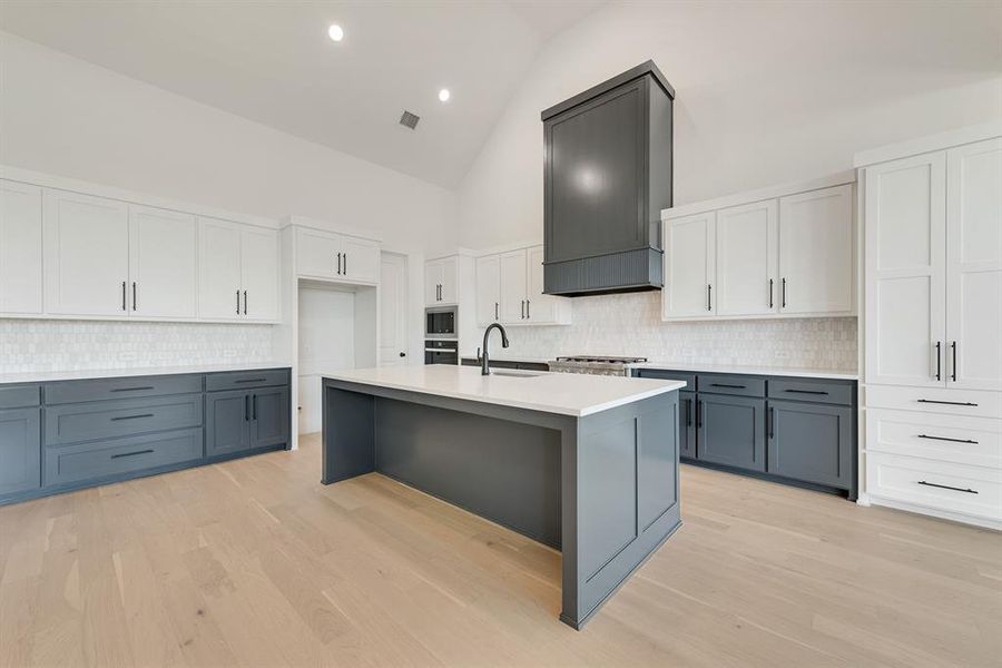 Kitchen with gray cabinets, tasteful backsplash, white cabinets, high vaulted ceiling, and light wood finished floors Kitchen with gray cabinets, tasteful backsplash, white cabinets, high vaulted ceiling, and light wood finished floors