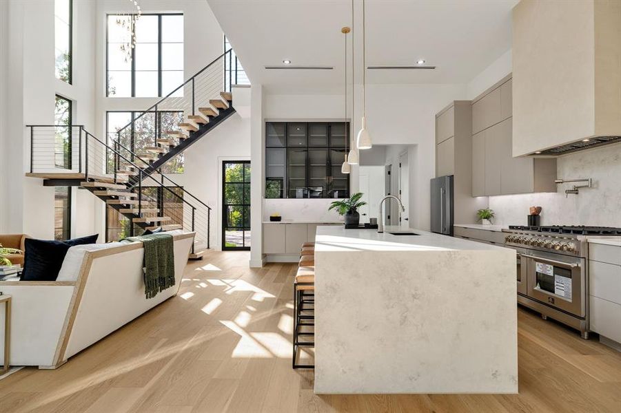 Open-concept living space featuring a floating staircase with cable railing, expansive multi-story windows, light wood-finish flooring, a waterfall island with an integrated sink, and stainless steel kitchen appliances