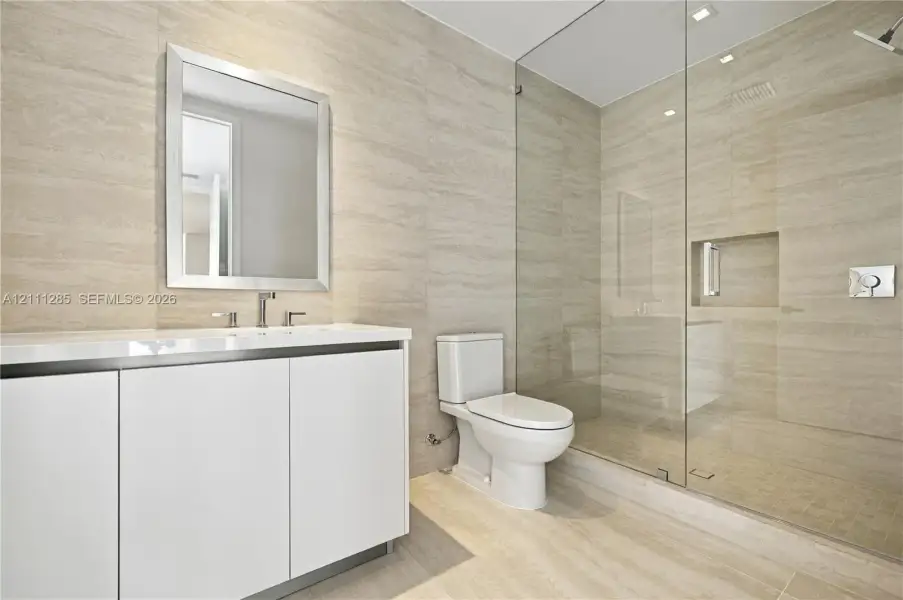 Wall-to-wall stone tile surfacing, walk-in glass-enclosed shower with a recessed shelf, white vanity with a rectangular sink, wall-mounted mirror, and a two-piece toilet Wall-to-wall stone tile surfacing, walk-in glass-enclosed shower with a recessed shelf, white vanity with a rectangular sink, wall-mounted mirror, and a two-piece toilet