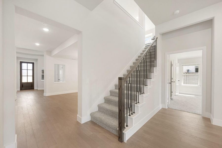 Open foyer with carpeted staircase, iron balusters, hardwood floors, and front entry door visible in background