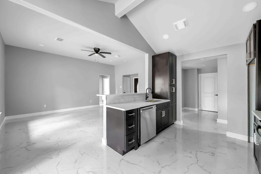 This photo showcases a modern, open-concept living space with a sleek kitchen featuring dark cabinets, a central island with a sink, and stainless steel appliances. The room has high ceilings, recessed lighting, and a ceiling fan, with light gray walls and glossy, marble-like flooring.