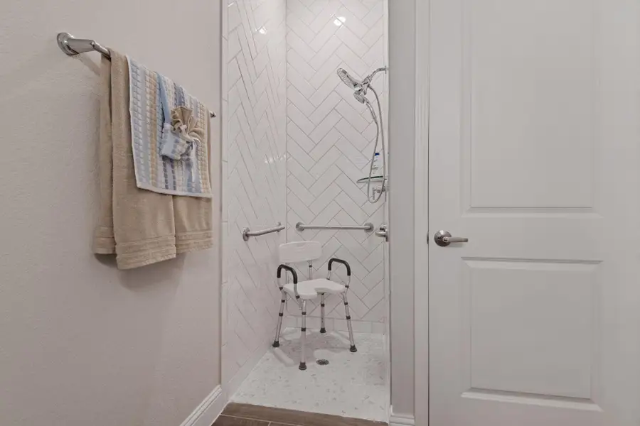 This photo shows a modern, ADA accessible bathroom with a walk-in shower. It features white herringbone tile, a handheld showerhead, and safety grab bars. A shower chair is present, and towels are hung neatly on a wall-mounted rack. This photo shows a modern, ADA accessible bathroom with a walk-in shower. It features white herringbone tile, a handheld showerhead, and safety grab bars. A shower chair is present, and towels are hung neatly on a wall-mounted rack.