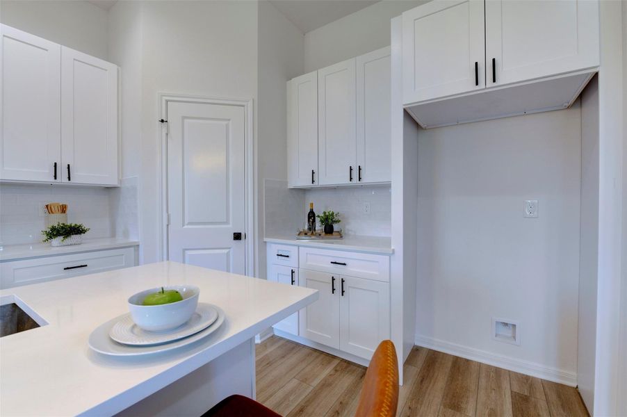 Kitchen storage wall showing floor-to-ceiling white shaker cabinets with matte black hardware, quartz countertop, subway tile backsplash, pantry door, and open appliance niche with overhead cabinet.