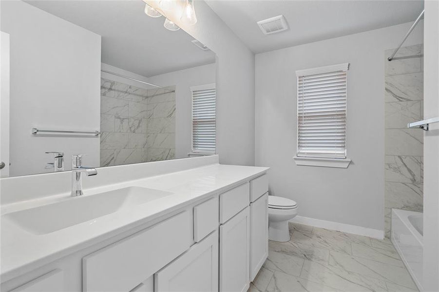 Secondary bath features tile flooring, bath/shower combo with tile surround, white stained wood cabinets, beautiful light countertops, mirror, dark, sleek fixtures and modern finishes.