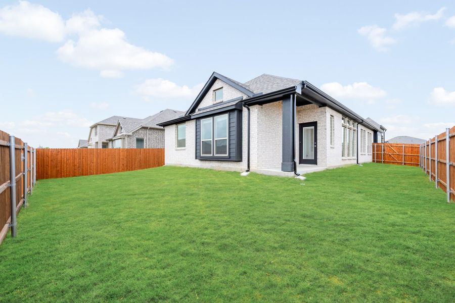 Backyard of single-story brick home with dark trim, large green lawn, and wood privacy fence