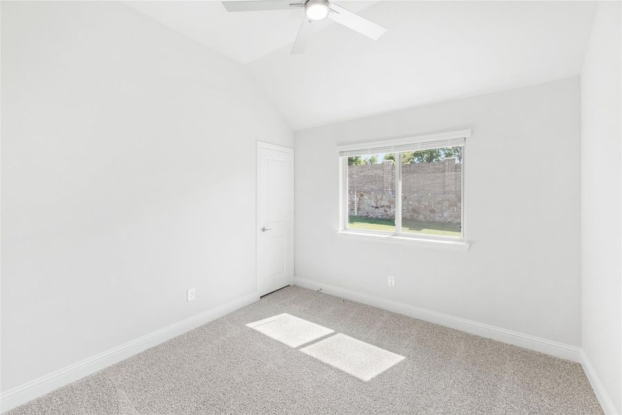 Empty bedroom with vaulted ceiling, ceiling fan, carpet flooring, and a window overlooking a backyard fence. Empty bedroom with vaulted ceiling, ceiling fan, carpet flooring, and a window overlooking a backyard fence.
