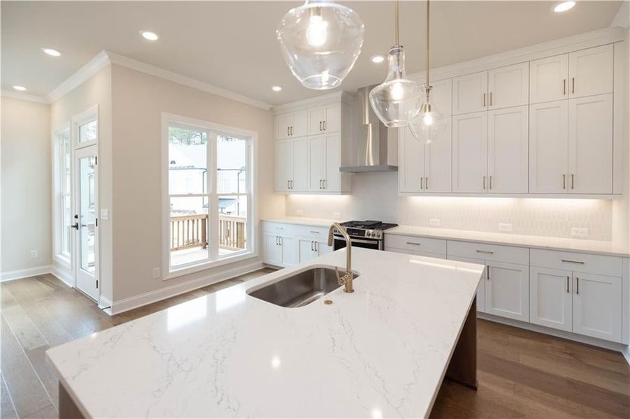 Kitchen with hanging light fixtures, stainless steel range with gas stovetop, an island with sink, wall chimney range hood, and white cabinets