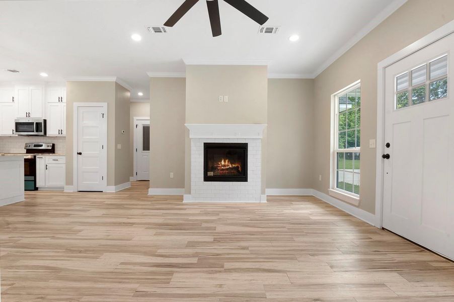 This bright, open-concept living space features a cozy fireplace, light wood flooring, and a ceiling fan. The adjacent kitchen has modern white cabinetry and stainless steel appliances, enhancing the home's contemporary appeal. Large windows and a front door with glass panels allow natural light to fill the room. This bright, open-concept living space features a cozy fireplace, light wood flooring, and a ceiling fan. The adjacent kitchen has modern white cabinetry and stainless steel appliances, enhancing the home's contemporary appeal. Large windows and a front door with glass panels allow natural light to fill the room.