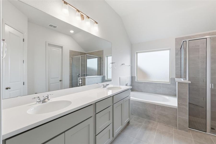 Bathroom featuring a dual vanity with light gray cabinetry and white countertops, a large soaking tub with gray tile surround, a separate glass-enclosed shower, and a vaulted ceiling Bathroom featuring a dual vanity with light gray cabinetry and white countertops, a large soaking tub with gray tile surround, a separate glass-enclosed shower, and a vaulted ceiling