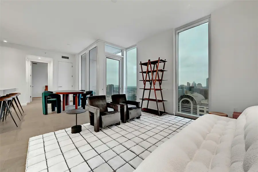 A minimalist design, featuring light-colored wood flooring arranged in a herringbone pattern. The room is brightly lit by natural light streaming in through large, floor-to-ceiling glass windows and doors that lead to a balcony. Furnishings provided by Scott + Cooner available for purchase separately. Photo is model unit #2406.