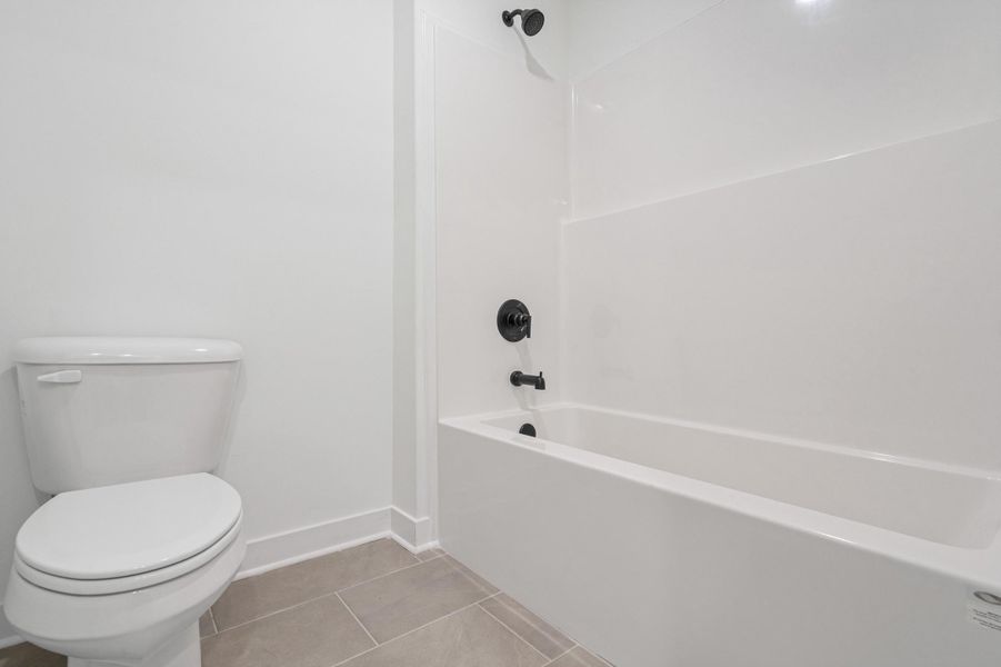 Bathroom featuring a white bathtub and shower combination with matte black fixtures, a white toilet, and rectangular floor tiles Bathroom featuring a white bathtub and shower combination with matte black fixtures, a white toilet, and rectangular floor tiles
