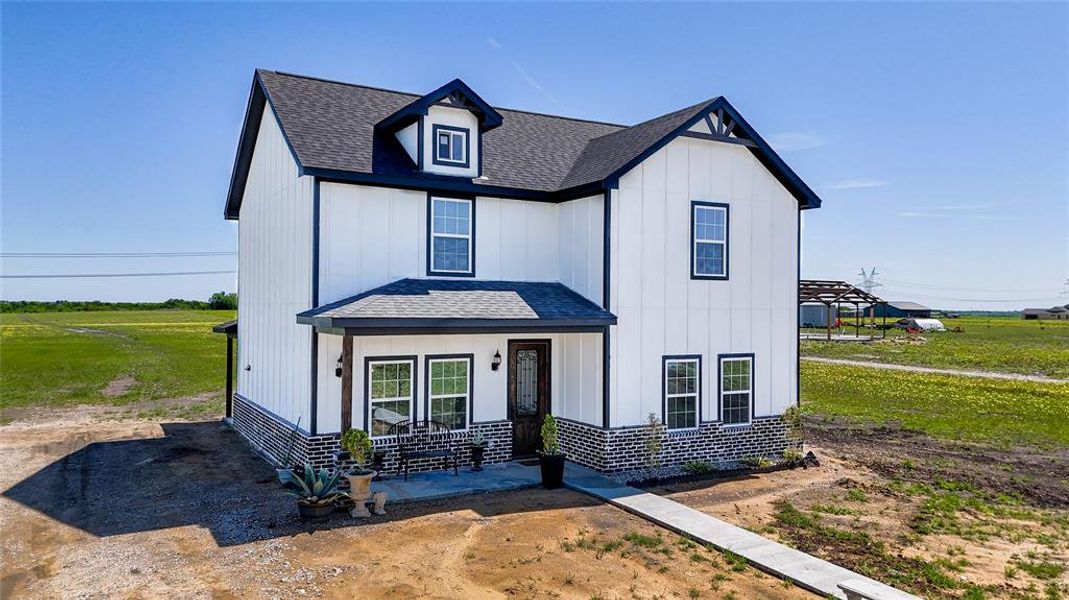 Two-story residence featuring white board-and-batten siding with dark trim accents, a dark asphalt shingle roof, a black brick foundation, and a covered front porch with exposed wood beams