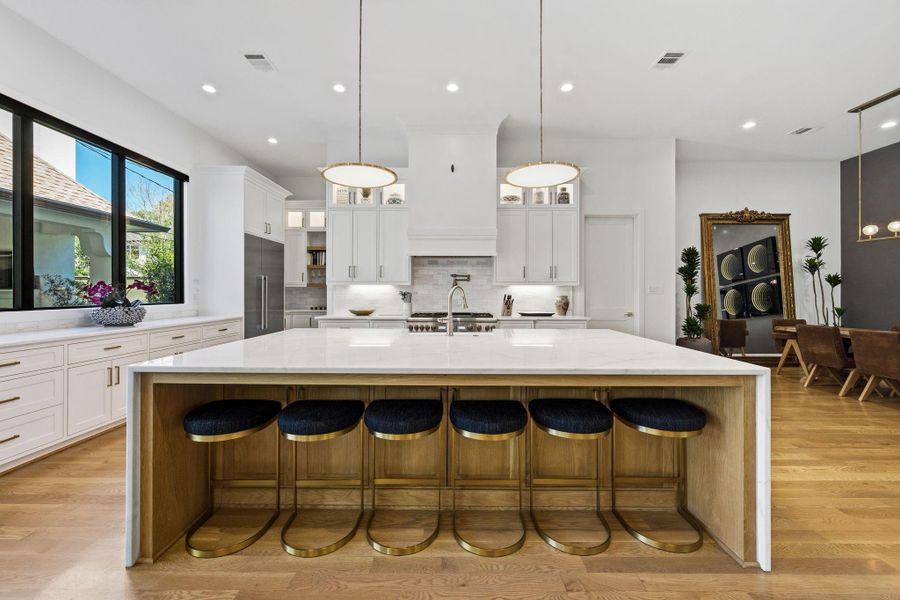 Striking chef’s kitchen anchored by an oversized waterfall-edge island with bar seating, creating a true centerpiece for gathering and entertaining. Features include wide-plank hardwood flooring, recessed lighting, designer pendant lighting, and custom cabinetry with under-cabinet lighting for both function and ambiance. Clean architectural lines and abundant natural light enhance the bright, refined aesthetic.