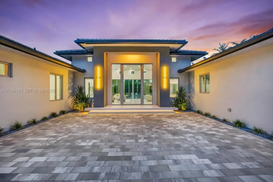 Modern stucco exterior featuring a tiered roofline, expansive paver courtyard, floor-to-ceiling glass entryway, and wall-mounted linear lighting