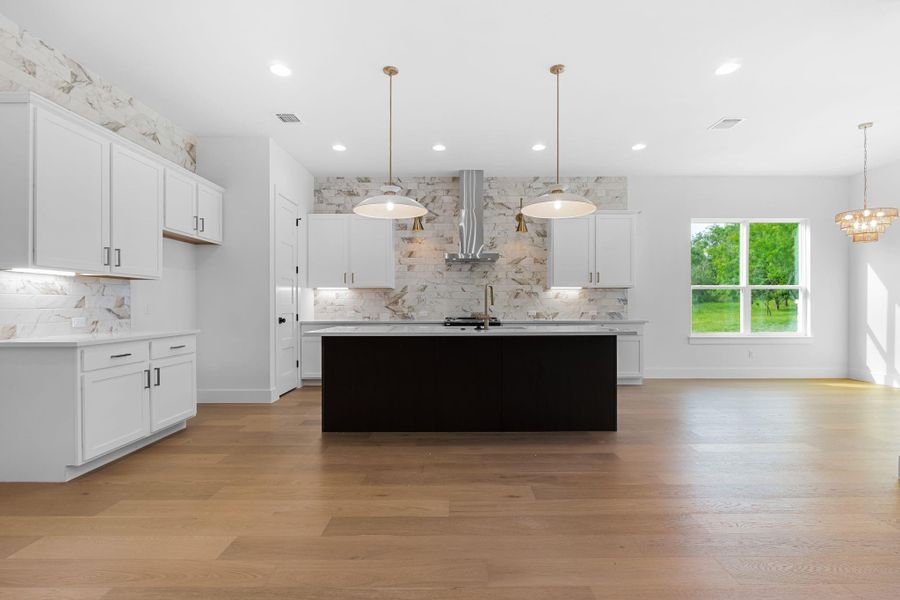Kitchen featuring hanging light fixtures, wall chimney exhaust hood, white cabinets, and a center island with sink Kitchen featuring hanging light fixtures, wall chimney exhaust hood, white cabinets, and a center island with sink