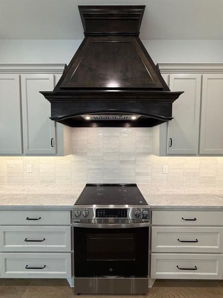 Kitchen with exhaust hood, range, light stone counters, white cabinetry, and decorative backsplash