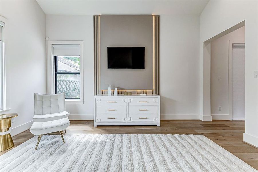 This room features modern decor with a sleek white chair with an ottoman, a stylish console, custom LED lighting, and a mounted TV. Natural light fills the space through a large window, highlighting the warm wood flooring and a plush area rug for a cozy and elegant atmosphere.