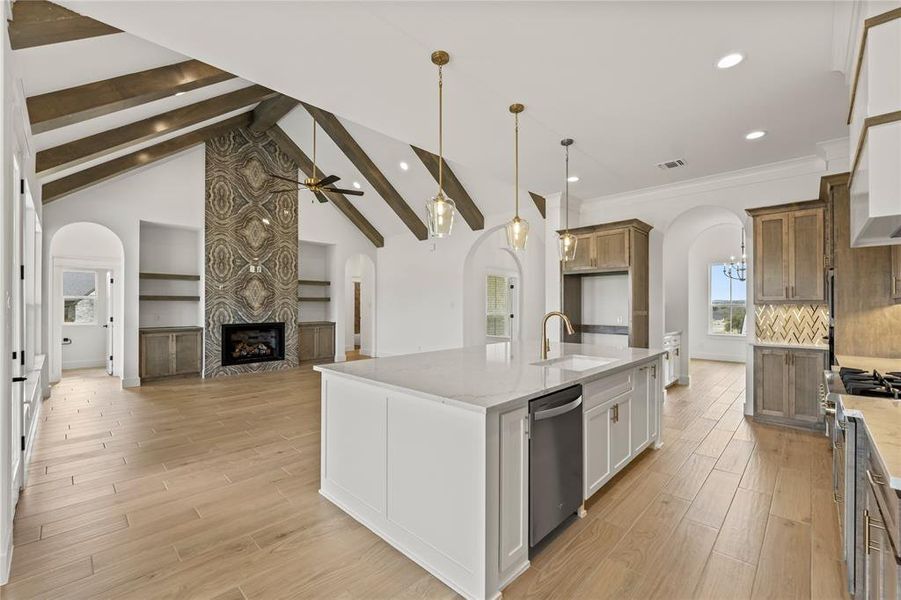 Expansive kitchen and living area featuring a central island with integrated sink, vaulted ceilings with wood beams, a fireplace with a decorative stone surround, wood-finish flooring, and arched doorways