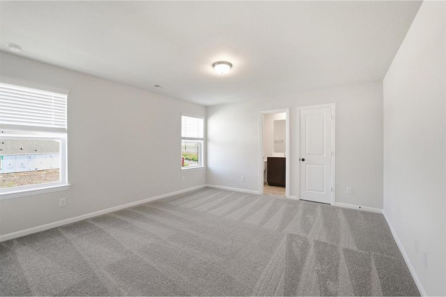 Spacious room featuring light gray carpeting, neutral wall paint, multiple windows with blinds, a ceiling-mounted light fixture, and an entry to a bathroom with a dark wood-finish vanity Spacious room featuring light gray carpeting, neutral wall paint, multiple windows with blinds, a ceiling-mounted light fixture, and an entry to a bathroom with a dark wood-finish vanity