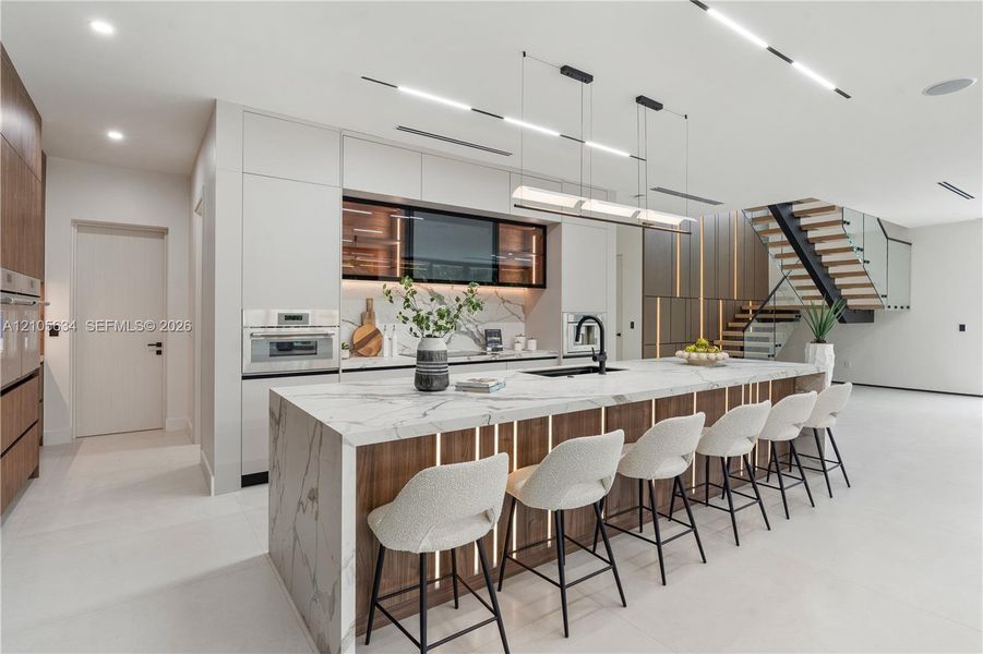 Modern kitchen featuring a waterfall-edge island with stone-finish surfaces and integrated wood-paneling, flat-panel cabinetry, built-in stainless steel appliances, and a staircase with a glass railing and wood treads