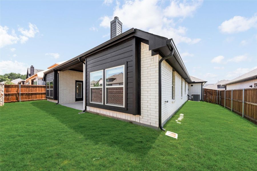Rear exterior of single-story home with dark siding, white brick, covered patio, and fenced backyard with green lawn