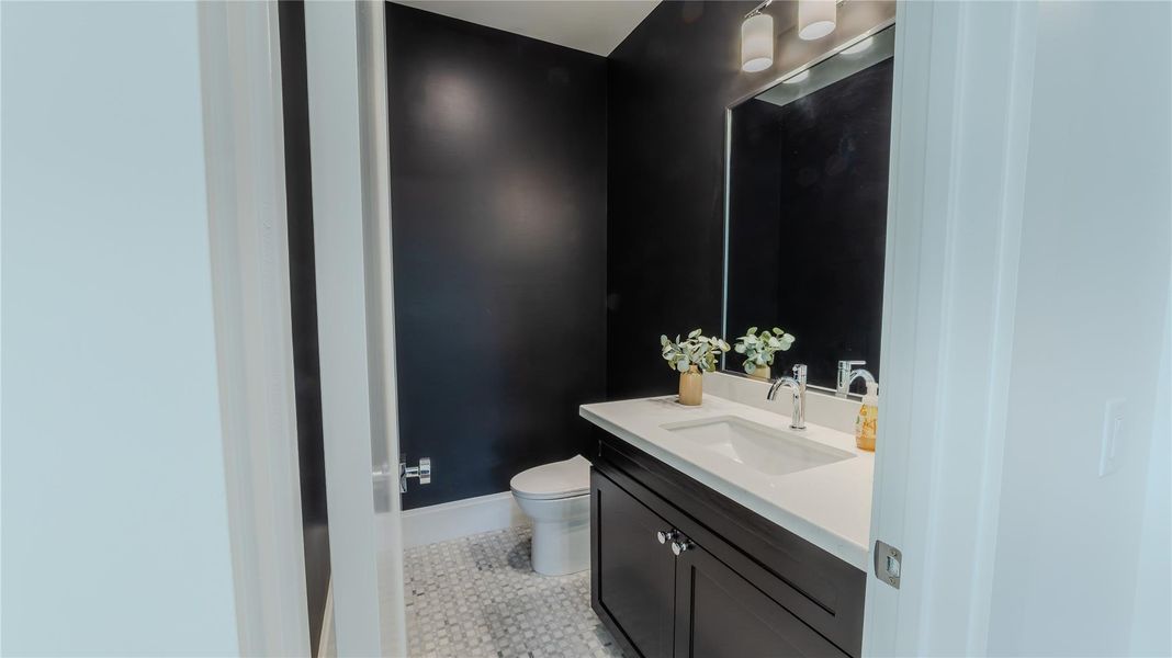 This bathroom features sleek black walls, a modern vanity with a white countertop, stylish lighting, and a decorative mirror. The floor has a classic tile pattern, and the space is accented with a small plant for a touch of greenery.