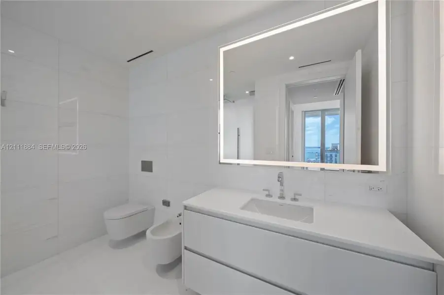 Modern bathroom featuring large-format stone-tile wall cladding, a wall-mounted toilet and bidet, a floating vanity with a stone countertop, and an integrated LED-backlit mirror Modern bathroom featuring large-format stone-tile wall cladding, a wall-mounted toilet and bidet, a floating vanity with a stone countertop, and an integrated LED-backlit mirror