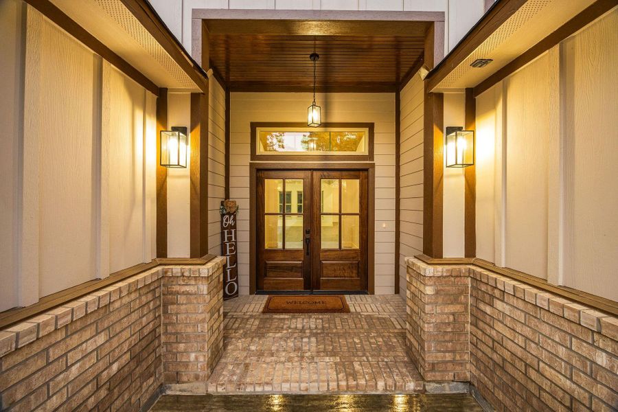 This inviting entrance features warm lighting, a wooden double door with glass panels. The space is framed by brick accents and paneled walls, creating a welcoming and elegant first impression.