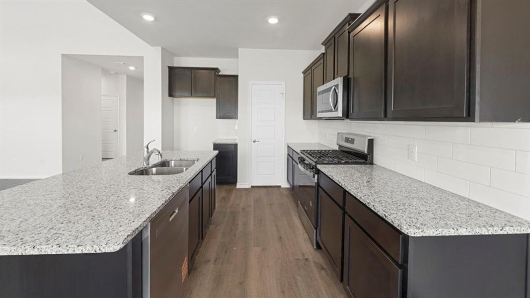 Open-concept kitchen featuring an oversized island with double basin sink, dark wood-finish cabinetry, stainless steel appliances, and wood-finish flooring Open-concept kitchen featuring an oversized island with double basin sink, dark wood-finish cabinetry, stainless steel appliances, and wood-finish flooring