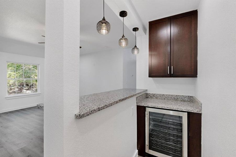 Indoor dry bar featuring wine cooler, dark brown cabinetry, light stone countertops, pendant lighting, and wood finished floors